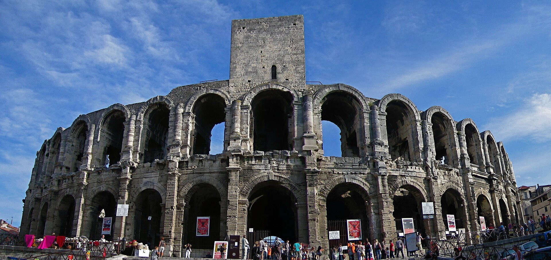 Amphithéâtre d’Arles, deux niveaux d’arcades, tour médiévale, public et affiches au pied des murs.