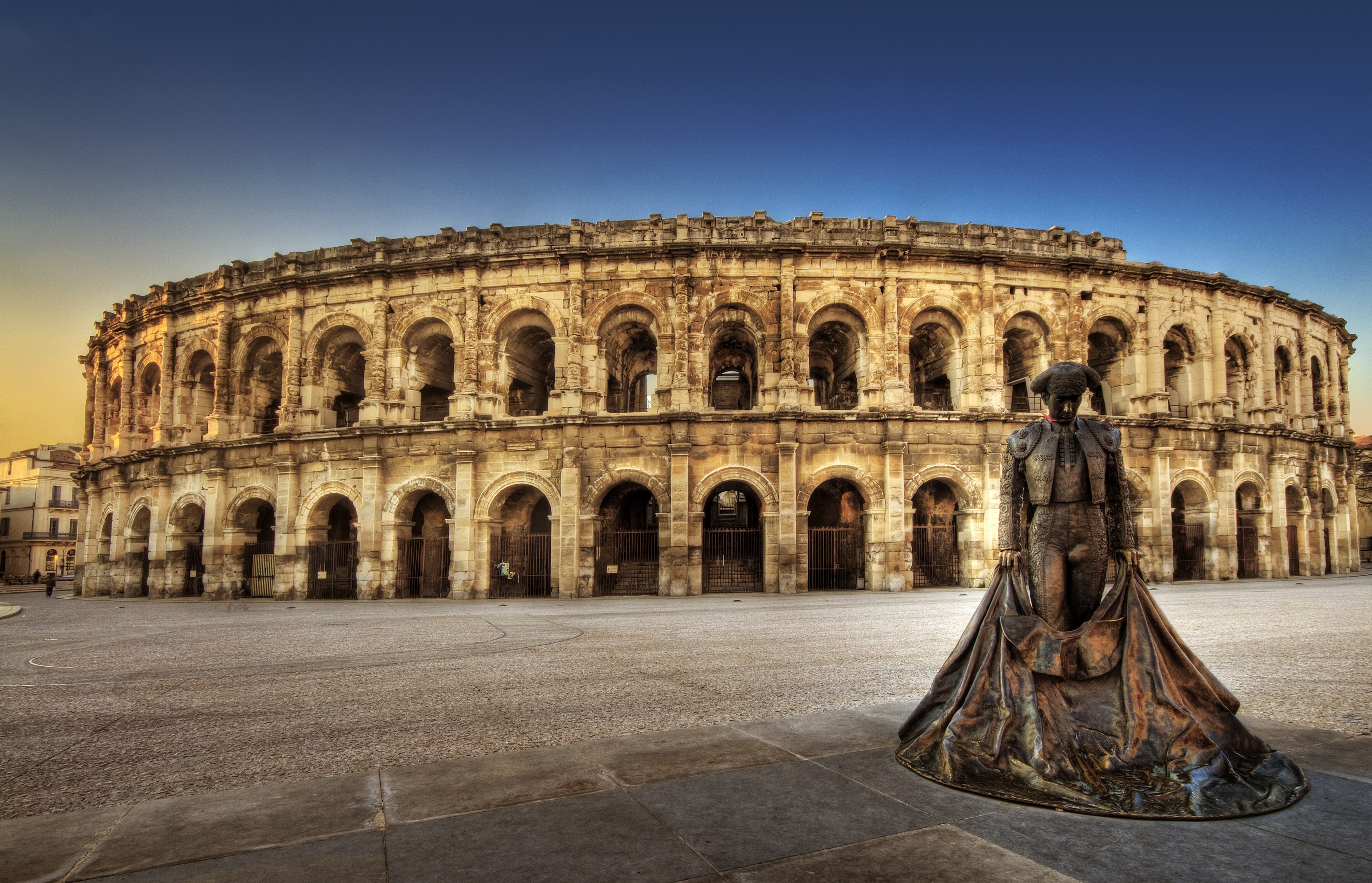 Arènes de Nîmes au crépuscule, statue de torero au premier plan, parvis vide.
